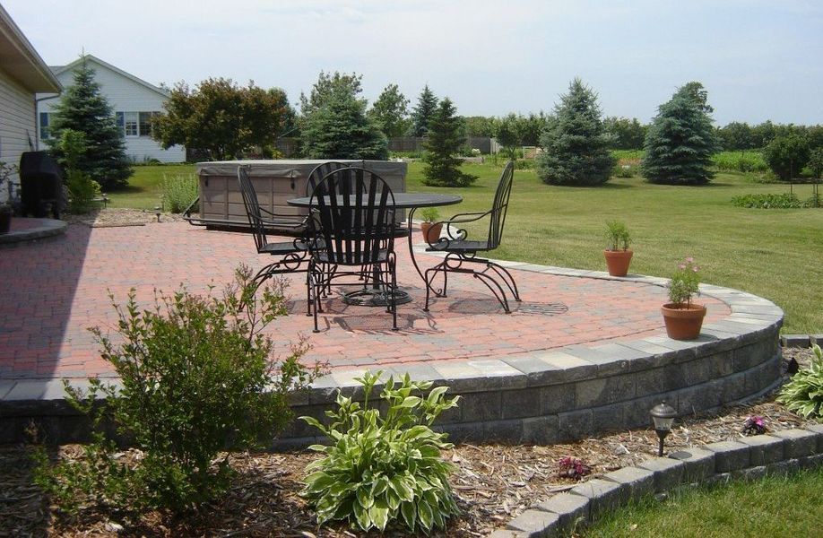 Patio with brick pavers, table, chairs, hot tub, and landscaping in a yard.
