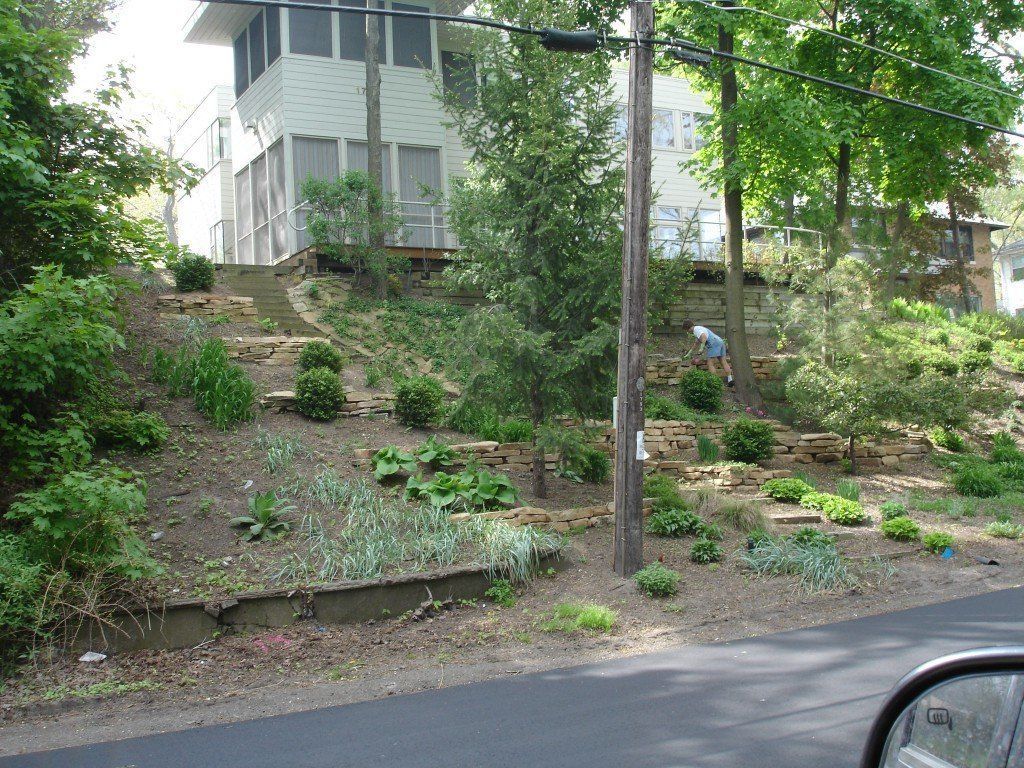 House with a tiered, landscaped front yard with stone retaining walls, a person working near the top.
