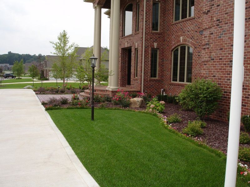 Lawn and flowerbed in front of a brick house with columns, a sidewalk on the left and a lamp post.