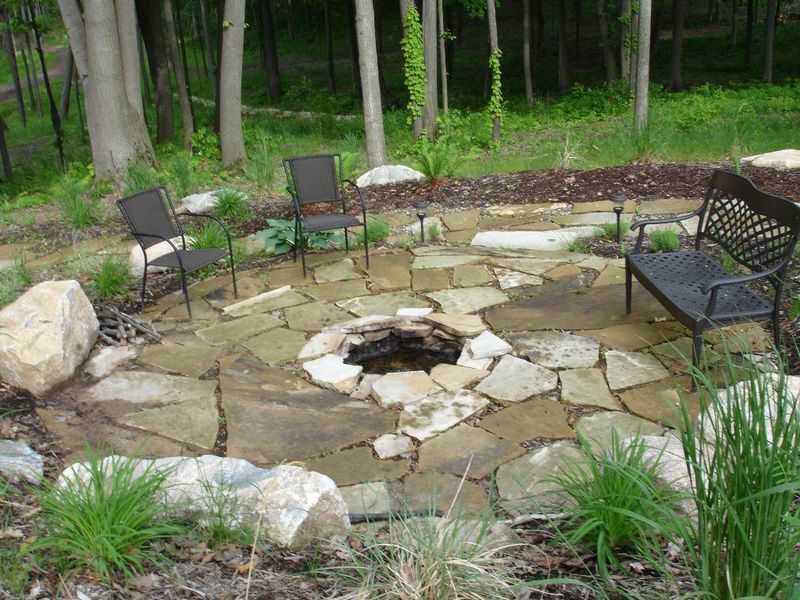 Stone patio with fire pit, seating, and trees in background.