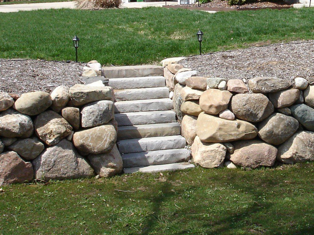 Stone steps between two stone walls in a yard, leading up to a mulched area with grass and small lights.