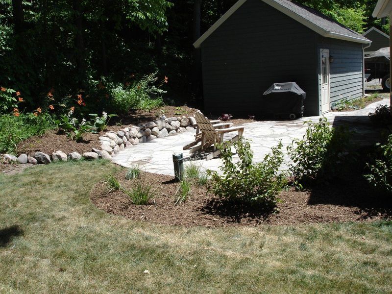 Stone patio with wooden chairs, next to a small gray shed, surrounded by garden beds and lawn.