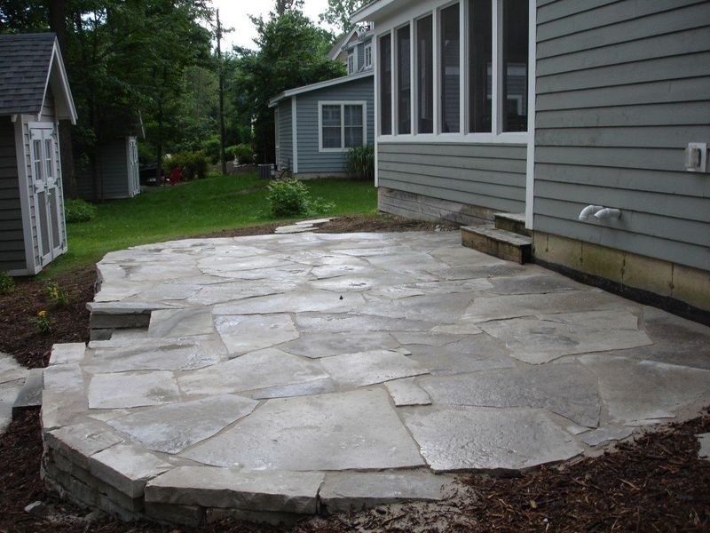 Stone patio next to a gray house with a screened porch, surrounded by mulch and grass.