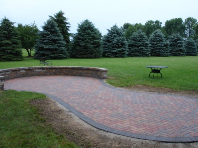 Brick patio with a curved stone wall, set in a grassy yard with evergreen trees in the background.