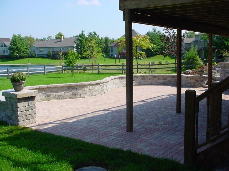 Brick patio with stone walls, overlooking a green lawn, pond, and houses under a clear blue sky.