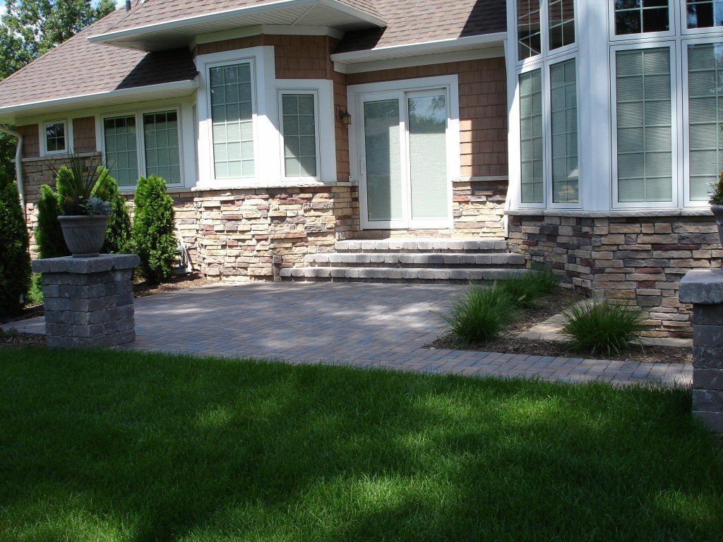 Stone patio and house entrance with steps and landscaping, with a grassy lawn in the foreground.