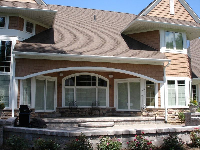 Large house with a brick patio, arched windows, and brown siding under a blue sky.