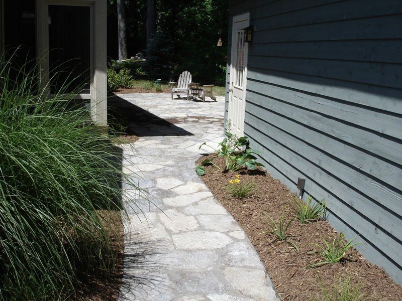 Stone path leads to patio with chair; side of a blue-gray house with landscaping on right and tall grass on left.