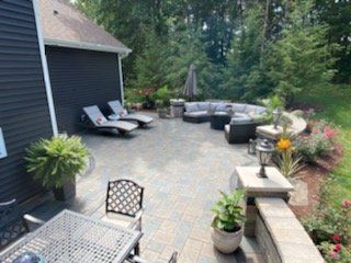 Patio with lounge chairs, curved sofa, dining table, and potted plants next to a house with a wooded background.