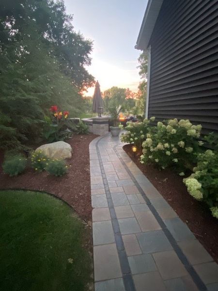 Brick pathway leads to an outdoor patio with plants and a dark house exterior at dusk.