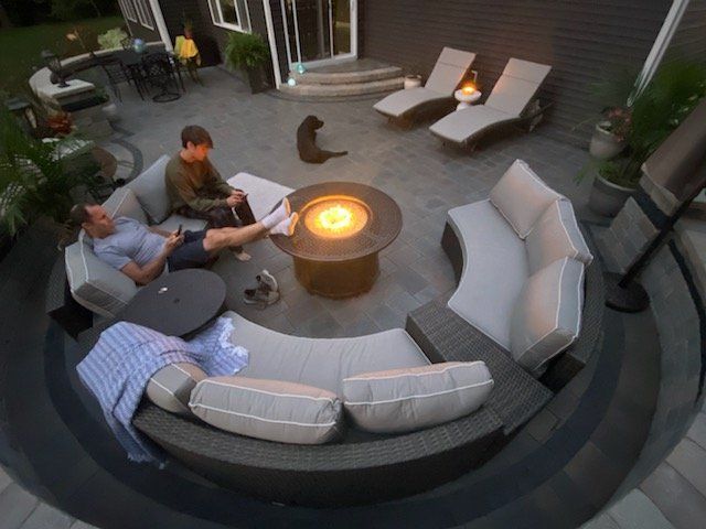 Two people relax on circular outdoor sofa around a fire pit with a cat nearby. Patio setting.