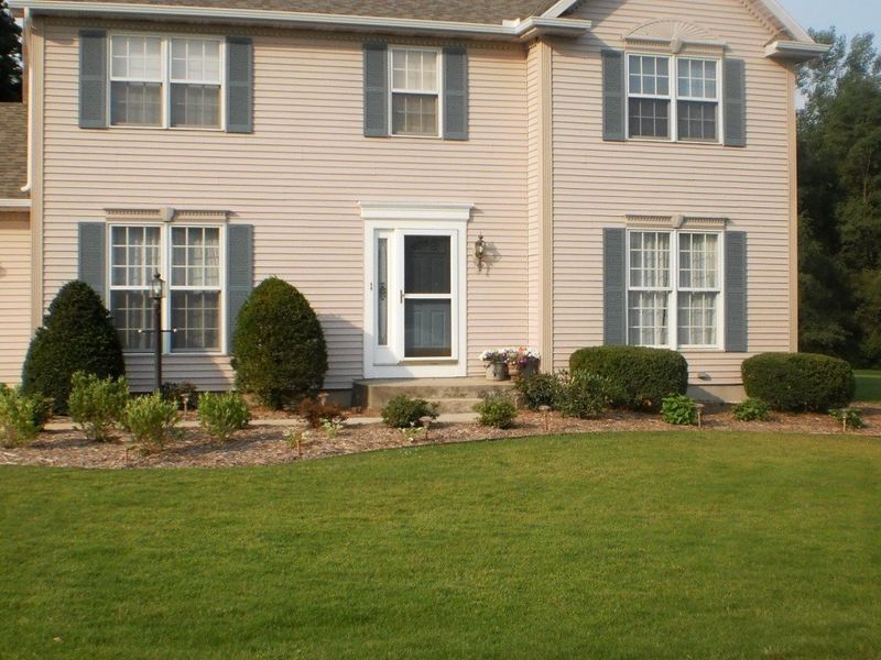 Beige two-story house with green shutters and lawn, and landscaping in front.