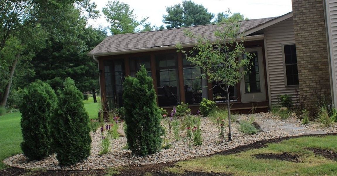 A house with a stone chimney and screen porch, surrounded by greenery and landscaping.