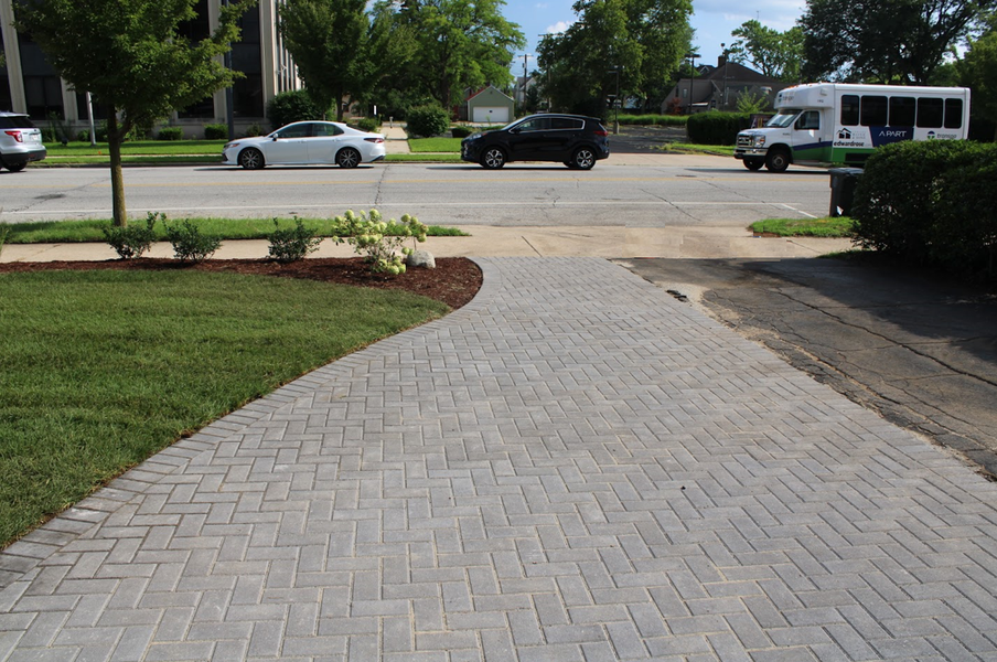 Brick path curves to a street with cars and a bus. Green lawn and trees on either side.