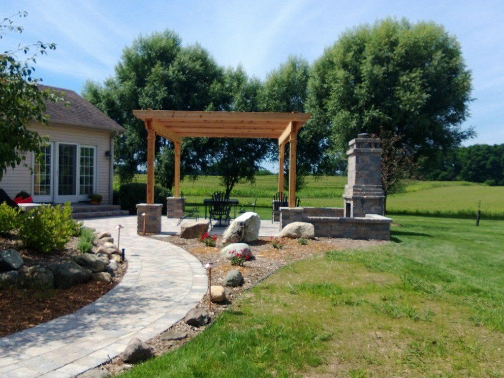 Backyard patio with pergola, fireplace, and stone pathway leading to the lawn and trees under a sunny sky.