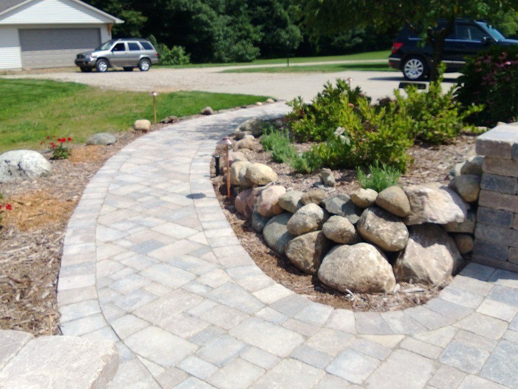 Stone walkway curves through landscaped yard with a rock retaining wall and parked cars.