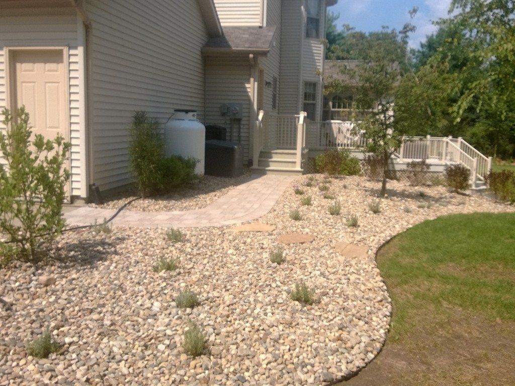 Exterior of a house with a stone pathway and landscaping. Beige siding, propane tank, and green grass are visible.