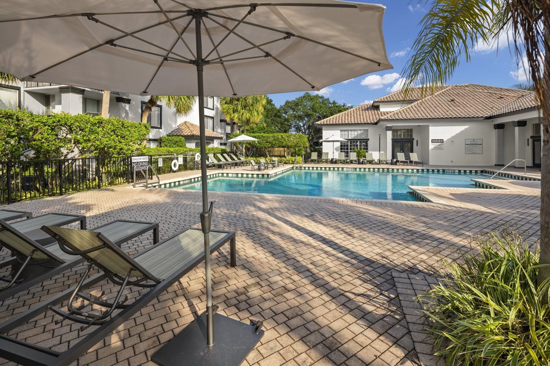 Poolside scene with lounge chairs, umbrella, and buildings in the background.