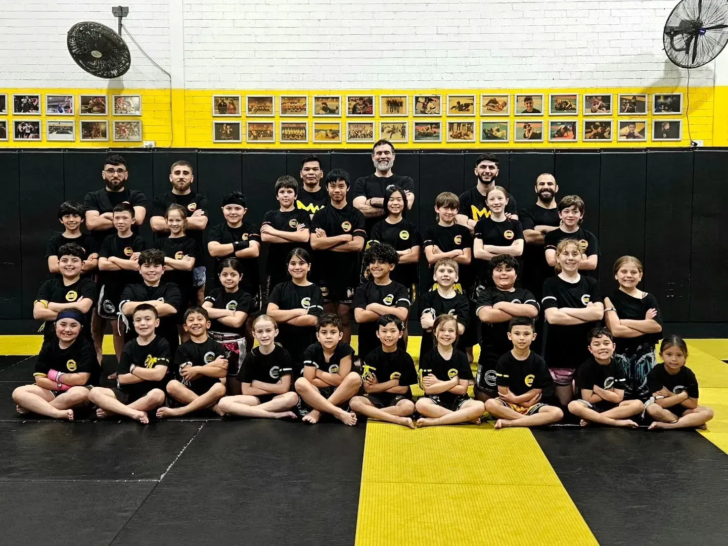 People in a martial arts gym stand around a yellow-and-black training mat, watching a sparring session.