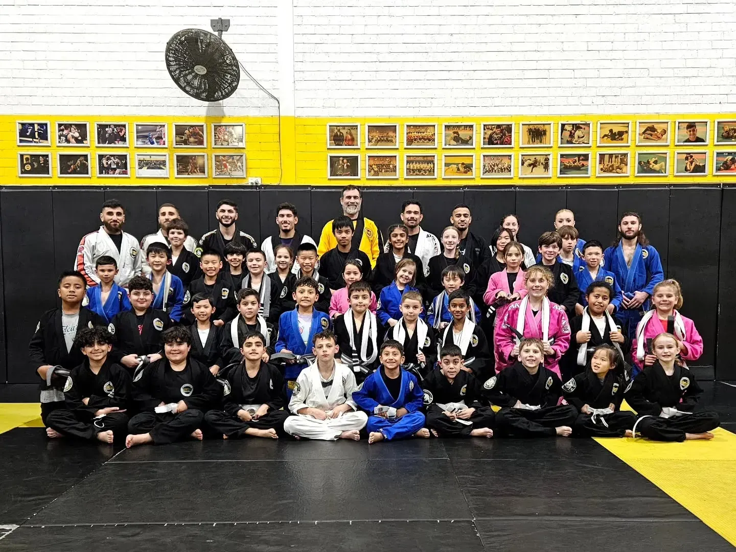 People standing in a circle around a black-and-yellow mat in a gym or martial arts studio.