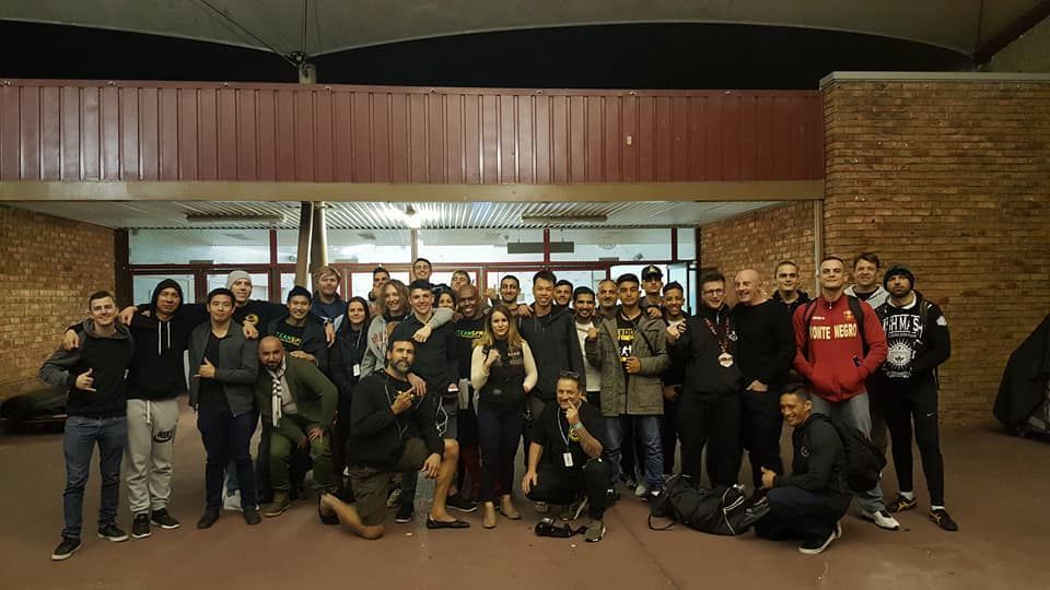 Large group posing outside a brick building at night, many holding medals or trophies, with one person crouching front right