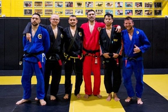 Six BJJ practitioners in gi stand on a mat in a gym with framed photos on the wall behind them.