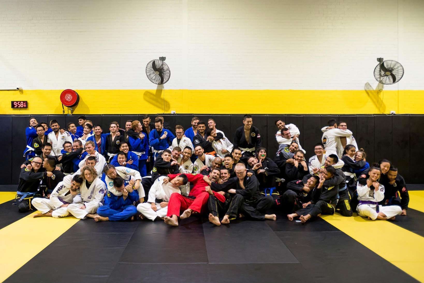 A large group in gi pose for a photo in a BJJ gym with mats, posters, and gym equipment behind.