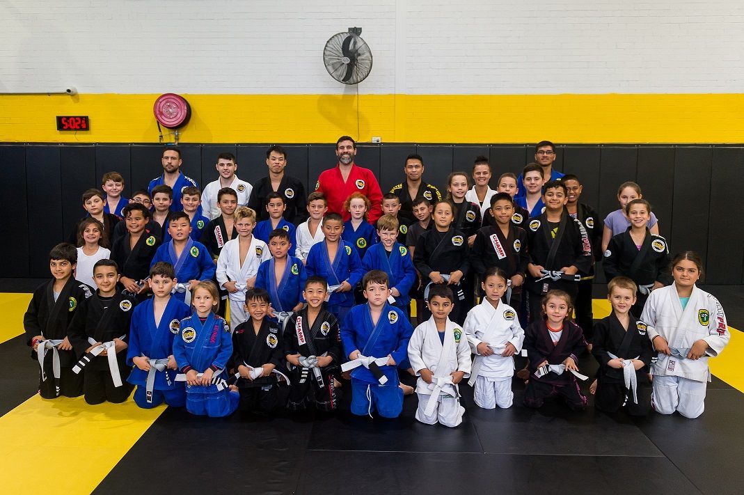 : A large group in gi pose for a photo in a BJJ gym with mats, posters, and gym equipment behind.
