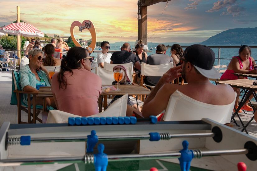 Persone in un bar sulla spiaggia con biliardino, cielo al tramonto e vista sul lago.