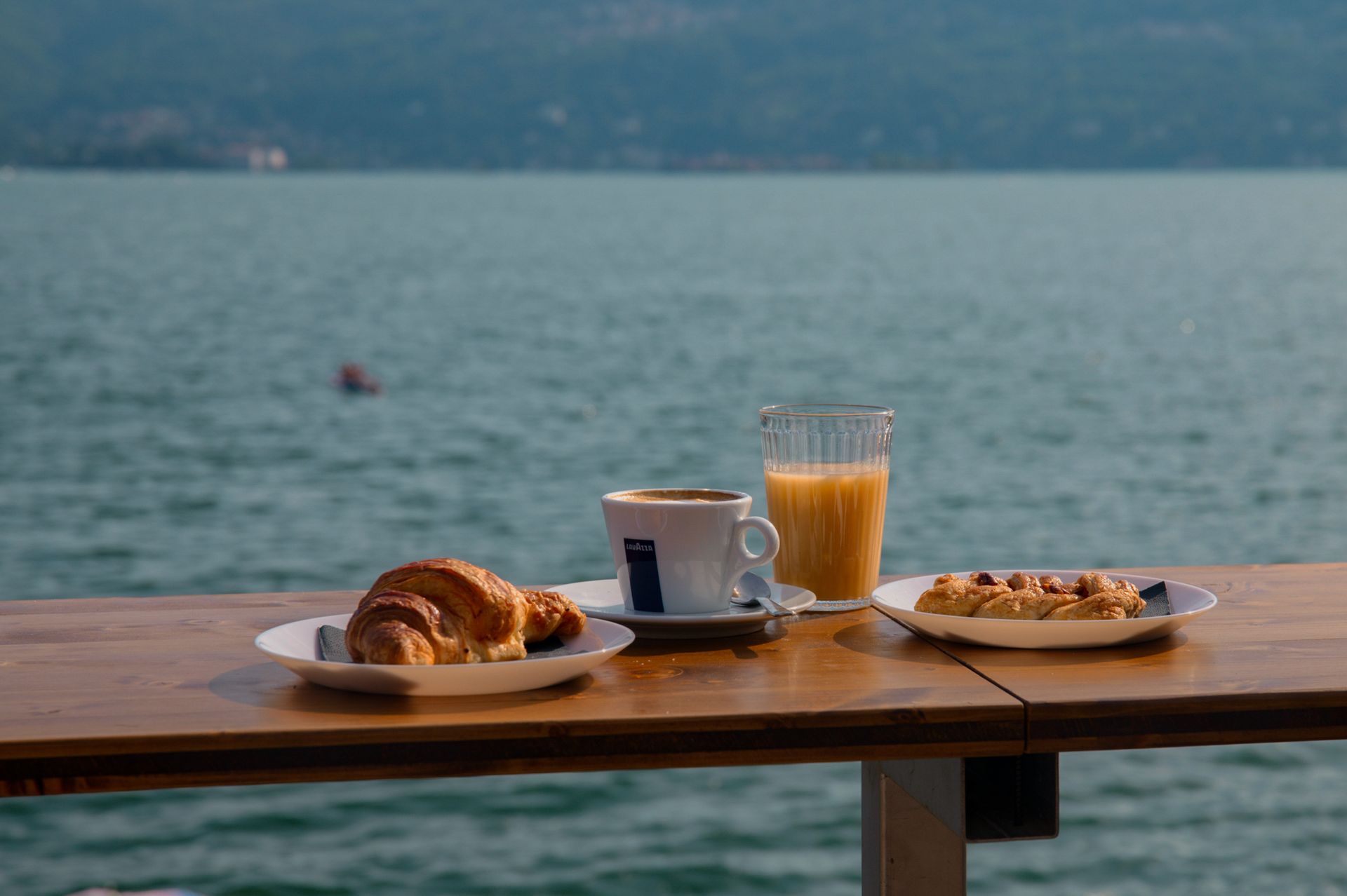 Colazione su un tavolo di legno con vista sul mare: croissant, caffè, succo di frutta e waffle.