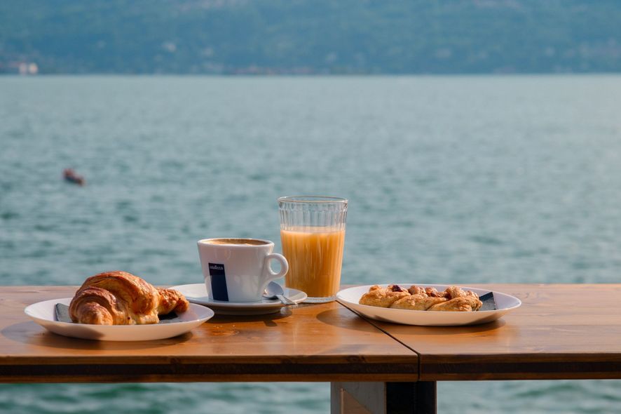 Tavolo per la colazione con croissant, pasticcini, caffè e succo d'arancia, con vista sul lago.