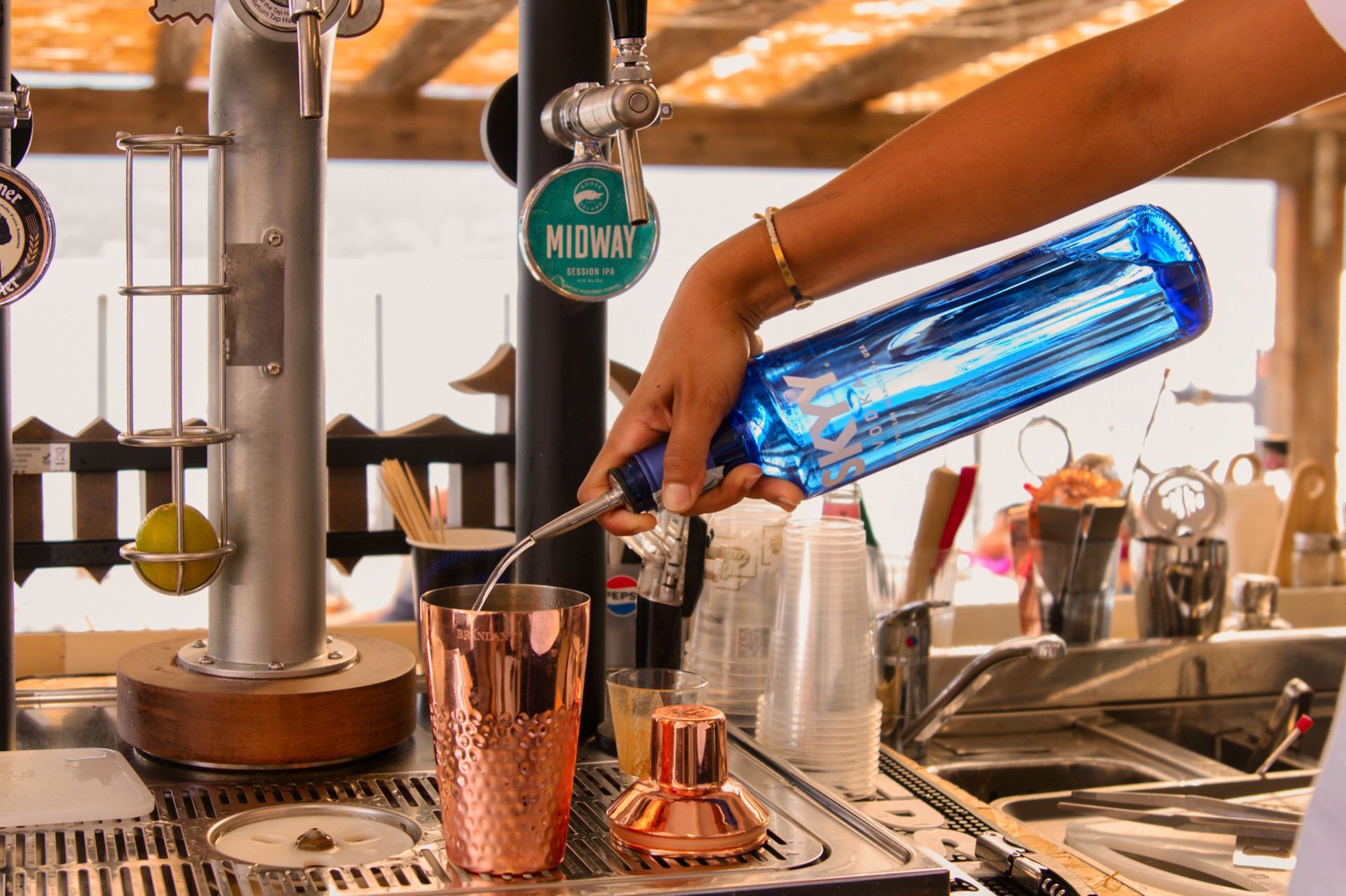 Barista che versa il liquido da una bottiglia blu in uno shaker di rame. Ambiente da bar sulla spiaggia.