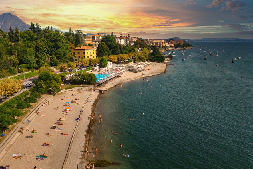Scena di spiaggia con persone che prendono il sole e nuotano. Edifici e alberi costeggiano la riva sotto un tramonto colorato.