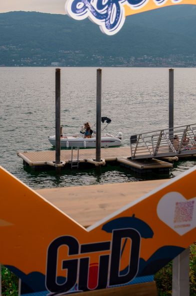 Persone su una piccola barca vicino a un molo di legno con le montagne sullo sfondo. Un cartello arancione e blu in primo piano.