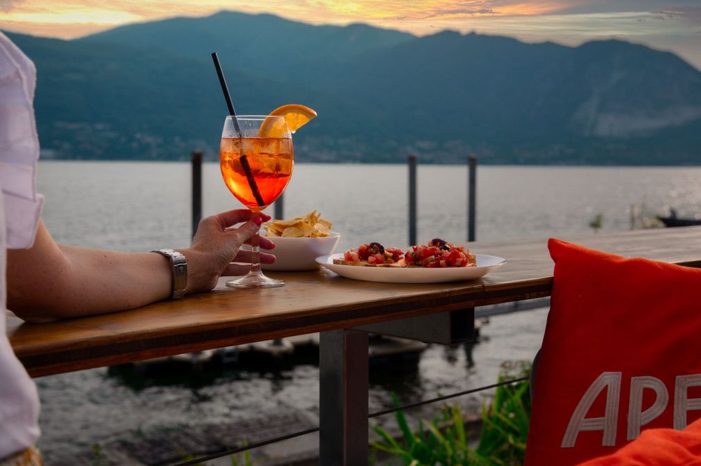 Persona che tiene in mano uno spritz Aperol in riva al lago al tramonto; piatto di cibo, patatine e cuscino con il marchio Aperol sul tavolo.