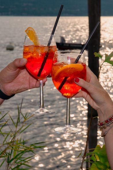 Due mani che brindano a un cocktail Aperol Spritz con un lago sullo sfondo.