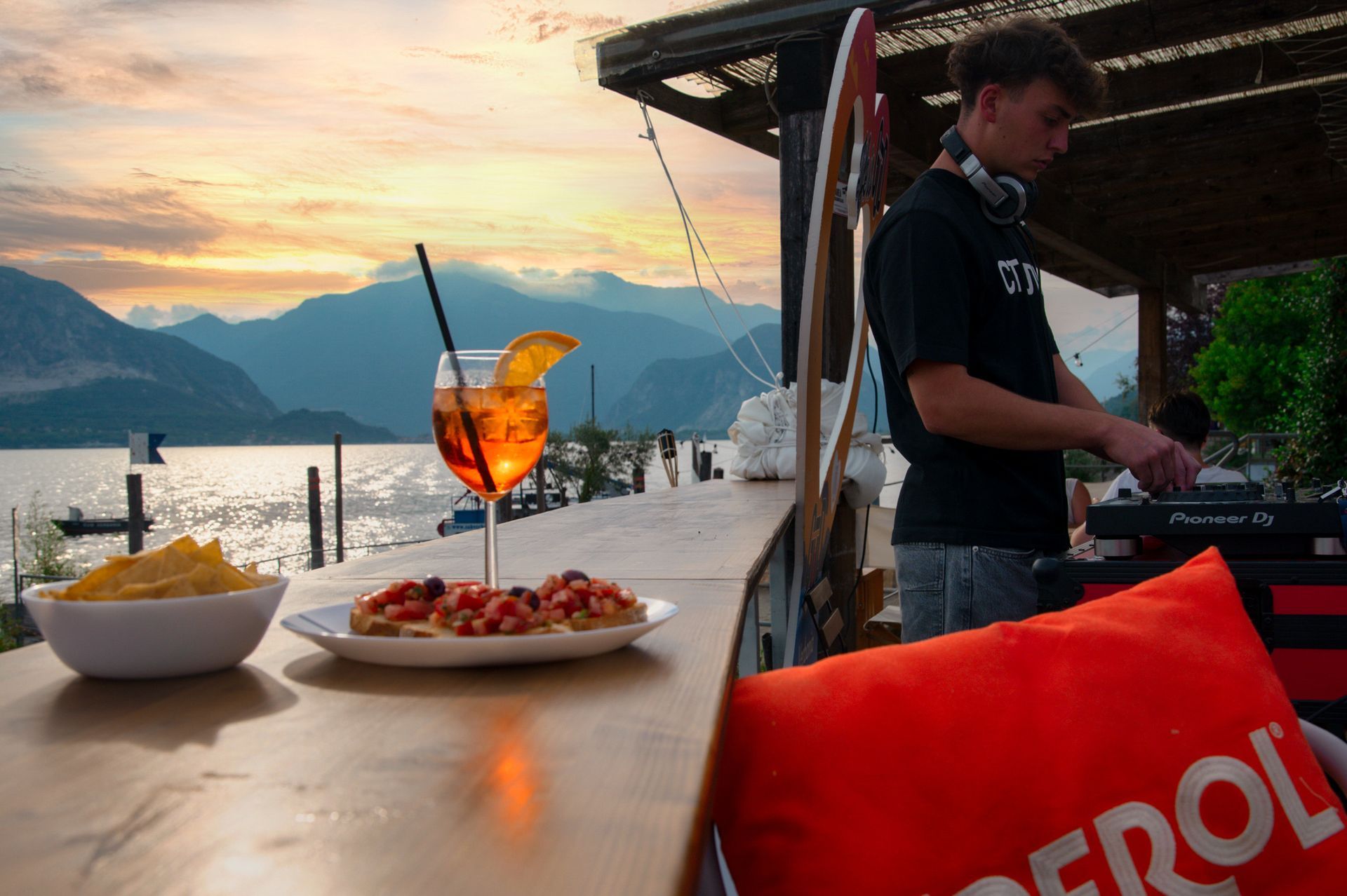 Bar al tramonto su un lago con cibo e bevande. DJ in sottofondo. Montagne in lontananza.