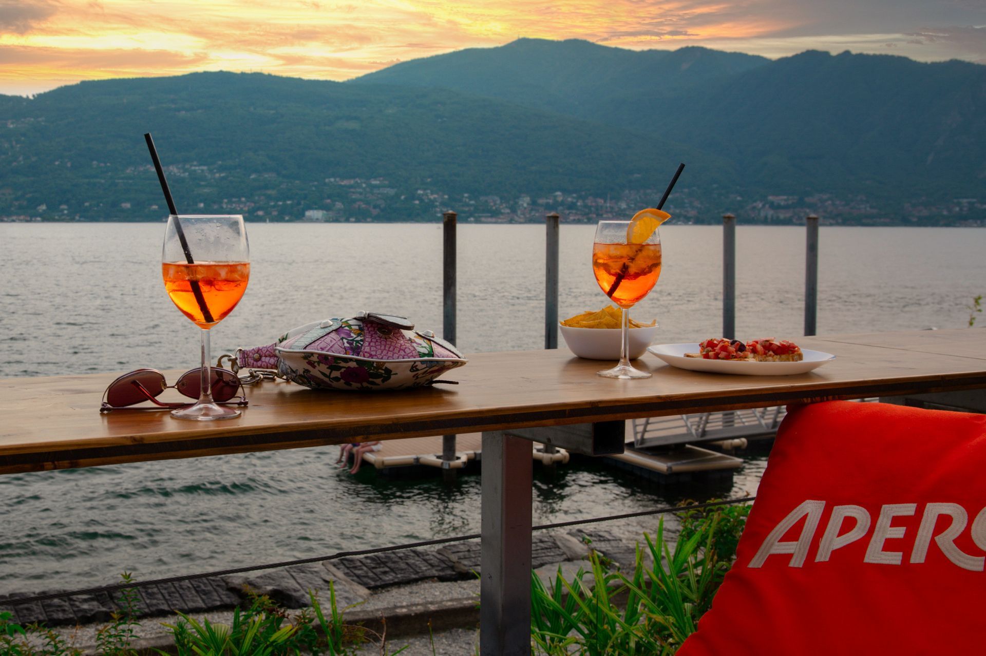 Due cocktail Aperol Spritz su un tavolo in riva al lago, con stuzzichini e le montagne sullo sfondo al tramonto.