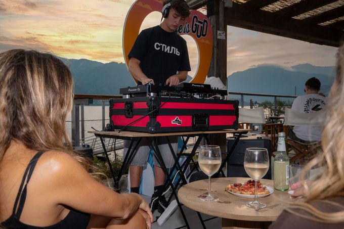 Un DJ suona musica su una terrazza con vista sul lago mentre due donne sorseggiano un drink, con il tramonto sullo sfondo.