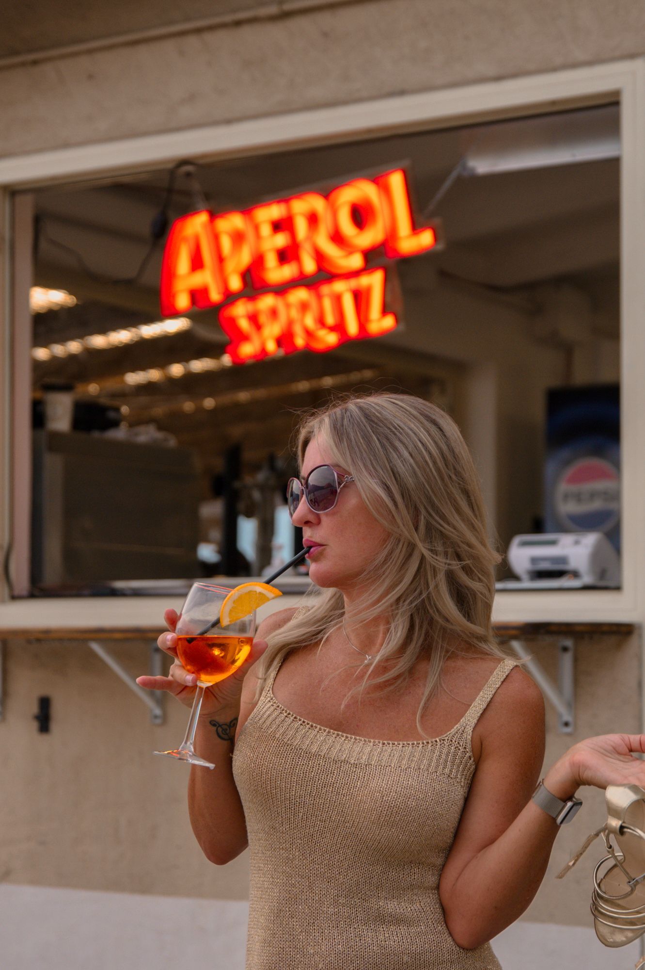 Una donna con occhiali da sole sorseggia un cocktail Aperol Spritz; sullo sfondo un'insegna al neon.
