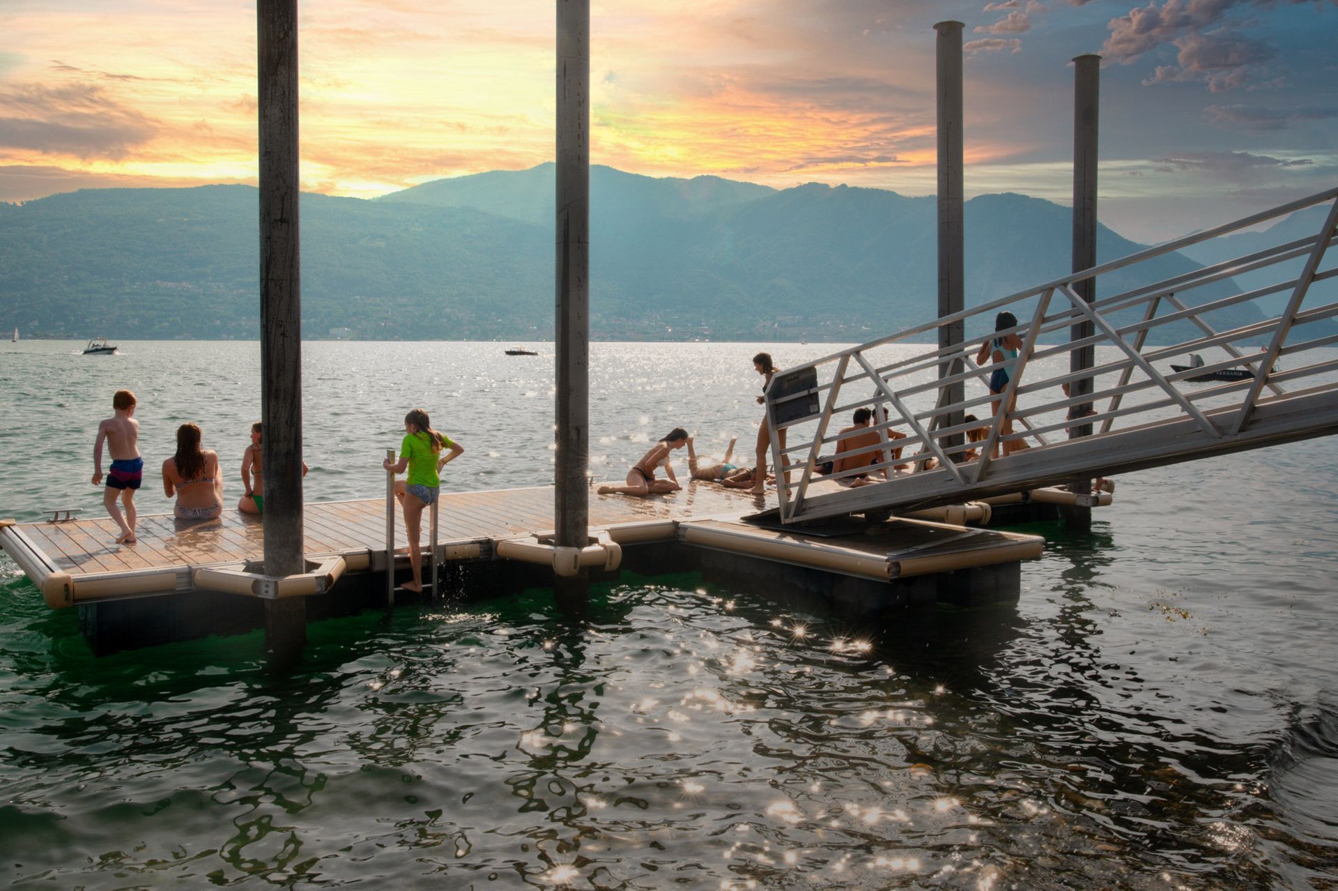 Coppia in un bar con vista sul lago e sulle montagne, che beve. Ombrellone a strisce rosse e bianche, tramonto.