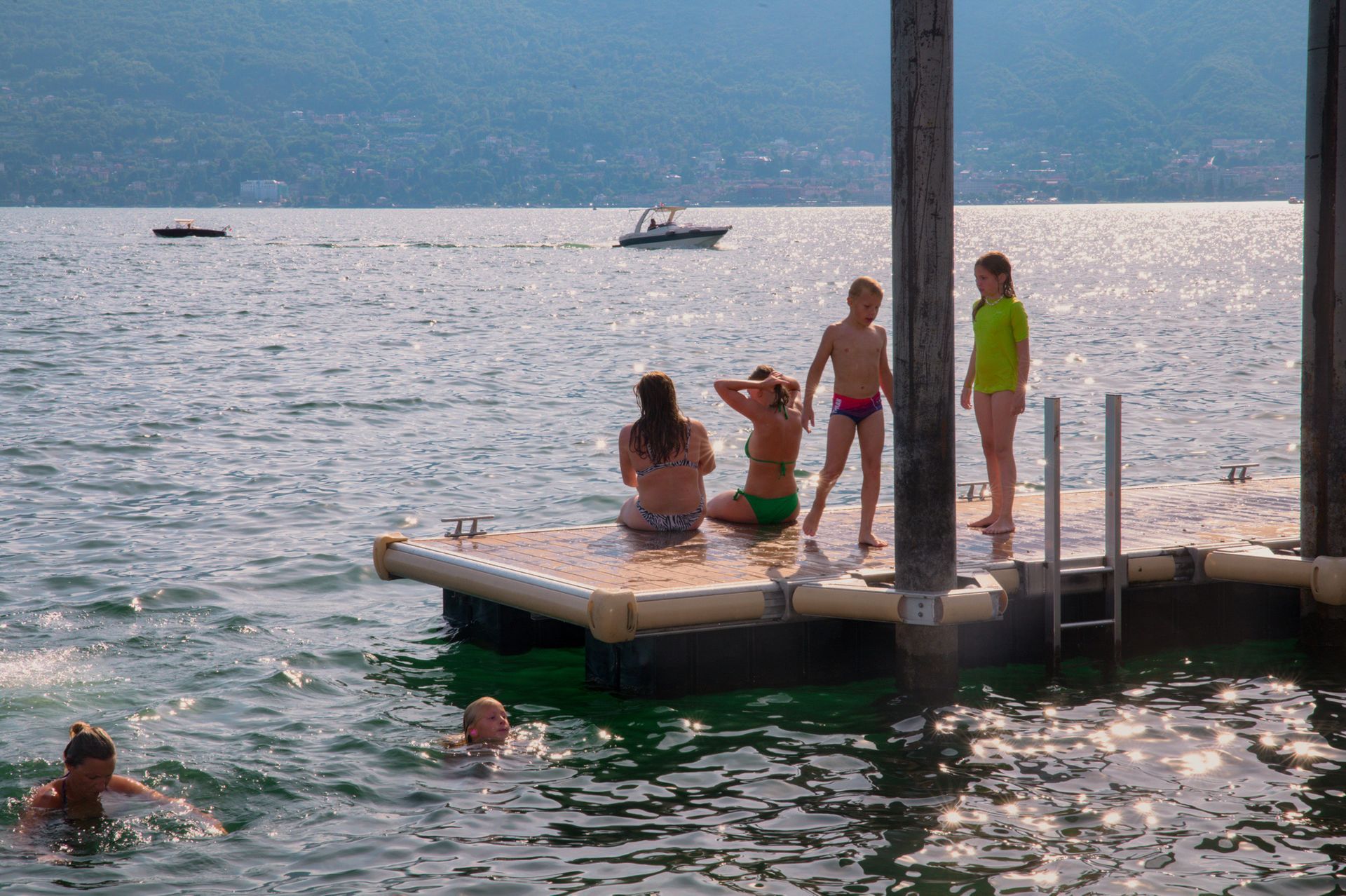 Persone che nuotano e si siedono su un molo in un lago in una giornata di sole, con le montagne sullo sfondo.