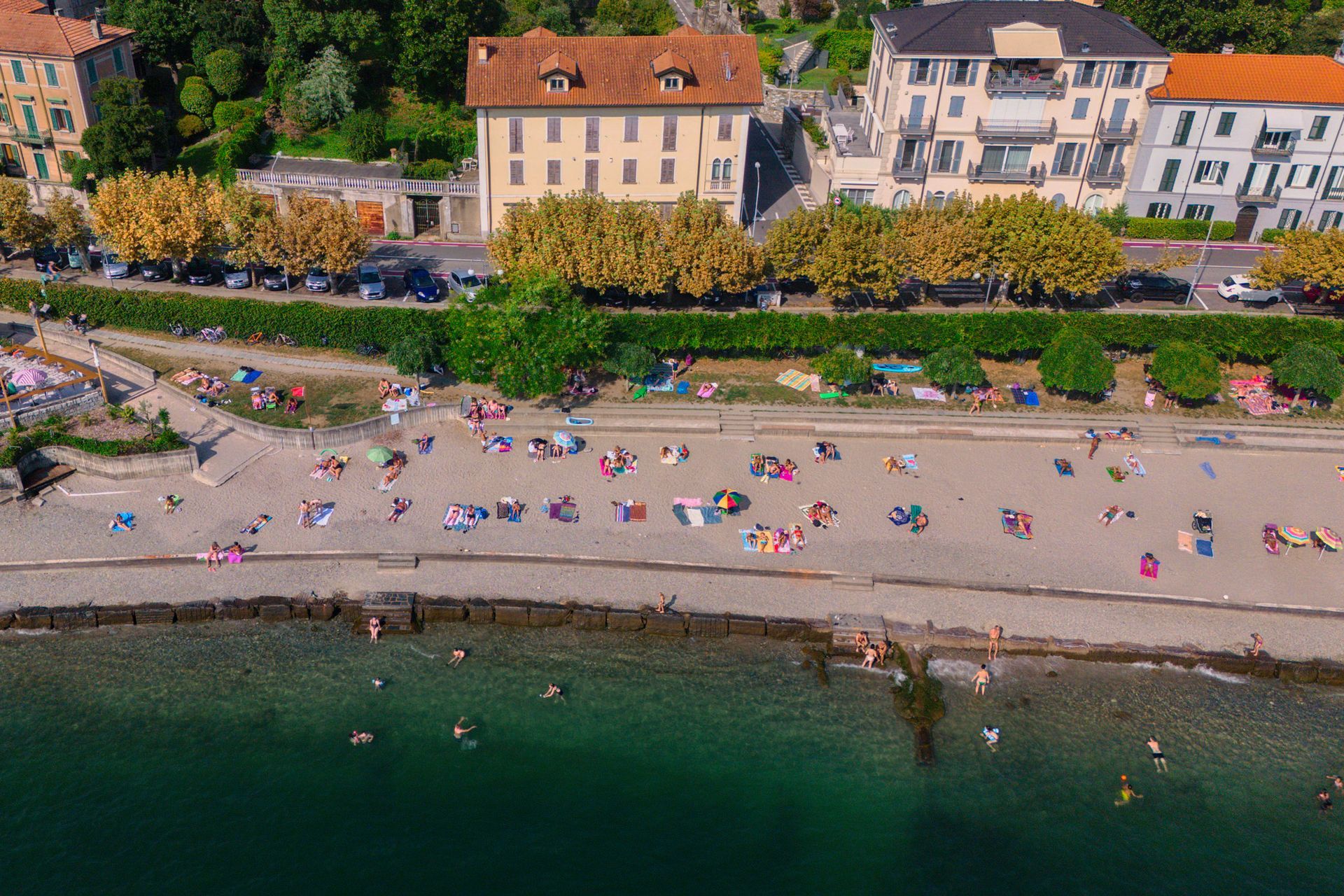 Scena di spiaggia con persone che prendono il sole, nuotano in acque turchesi, edifici lungo la riva.
