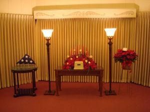 A memorial setup with a draped American flag, an urn centered on a table with red flowers, and two tall lamps.