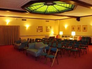 A meeting room with rows of chairs, a green stained-glass skylight, soft wall lighting, and a red carpet.