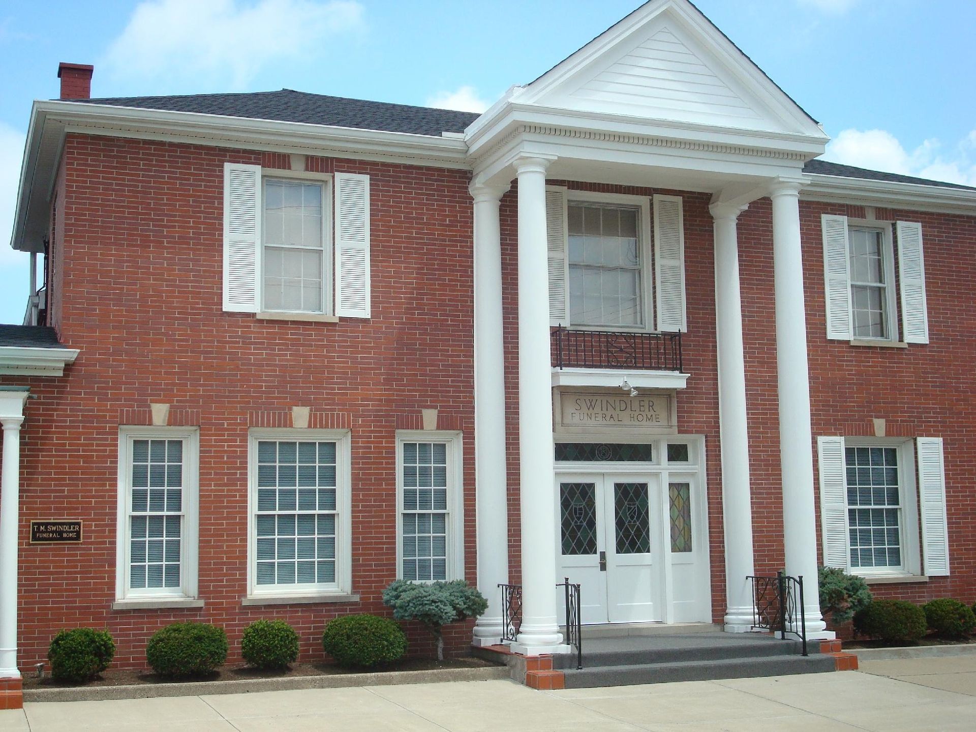 A red brick two-story building with white columns at the entrance and a small metal plaque on the side.
