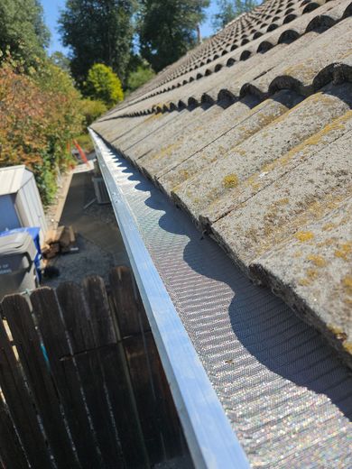 Gutter system with mesh screen along a roof. Gray roof tiles and a wooden fence are visible. Sunny outdoor setting.