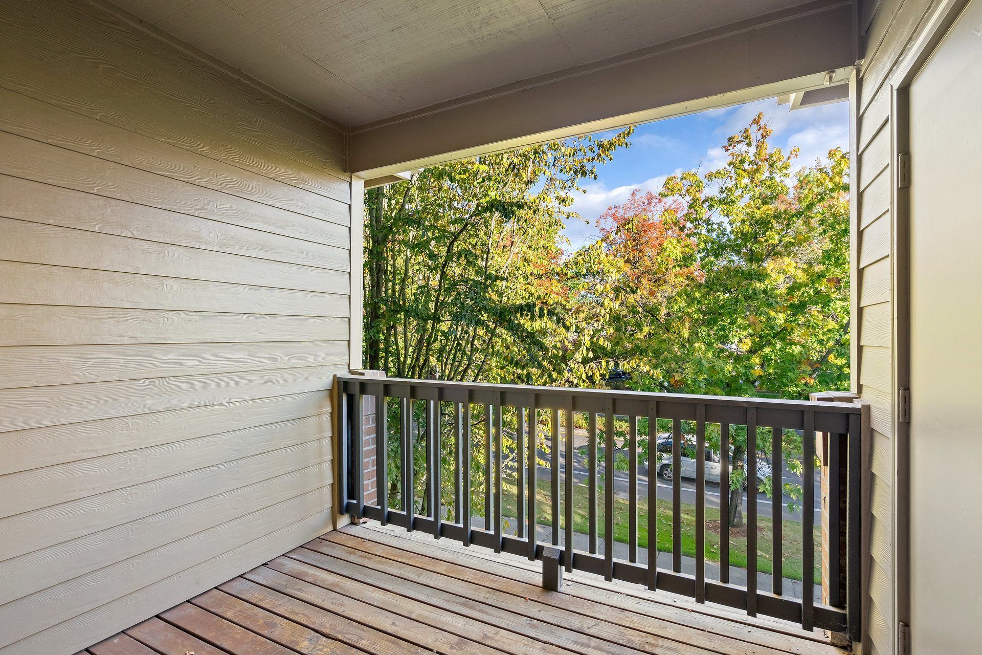 Balcony of an apartment with wood railing, beige siding, overlooking trees.