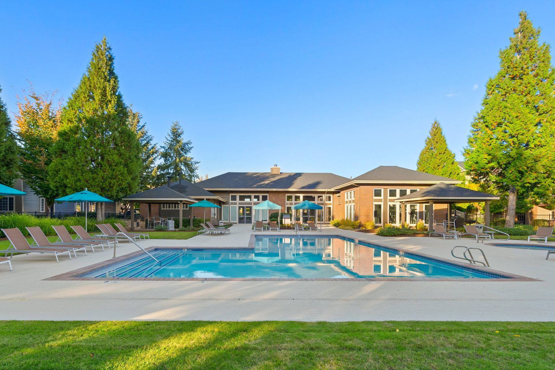 Outdoor community pool area with lounge chairs, teal umbrellas, and a clubhouse in the background.