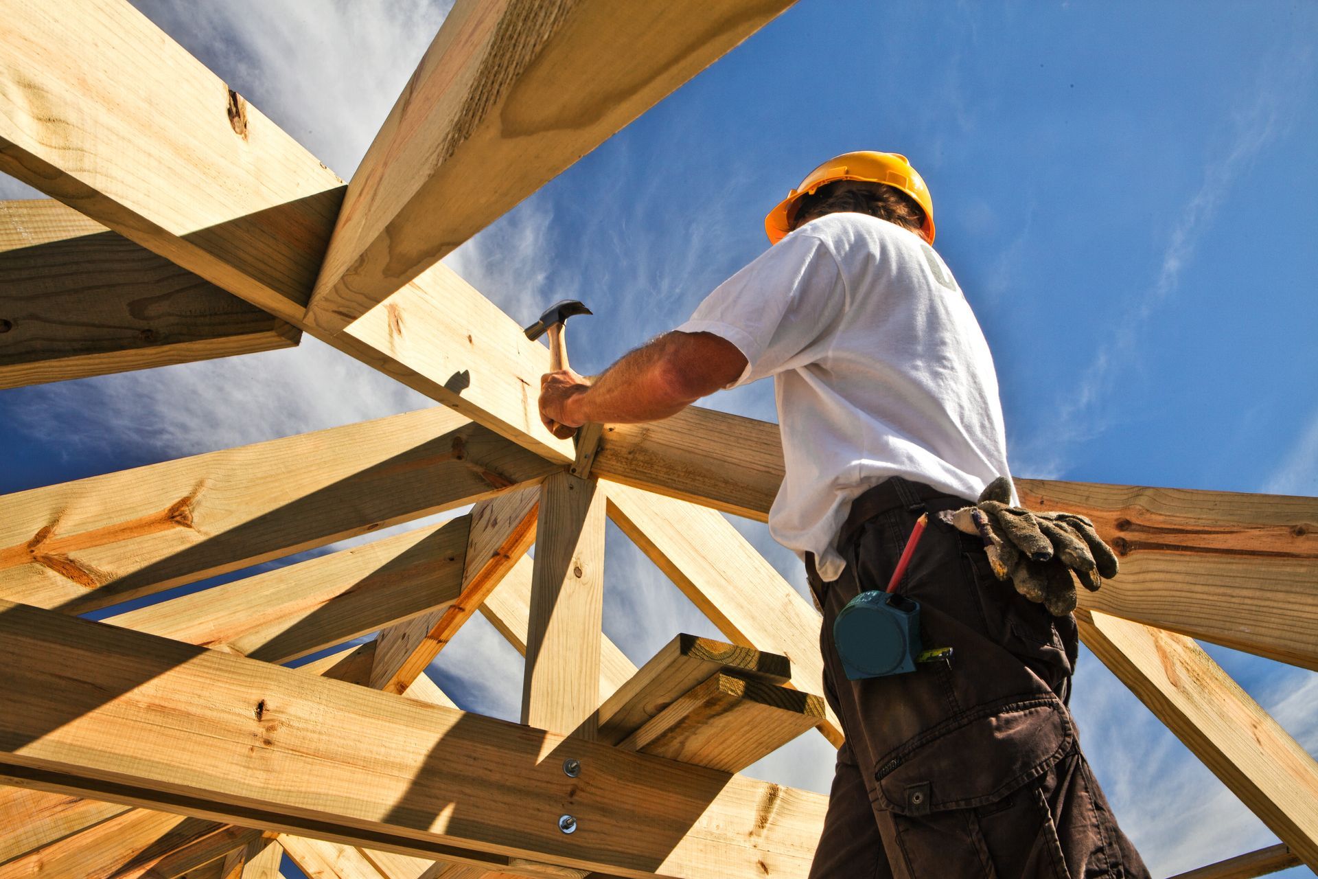 A construction worker in a yellow hard hat hammers a wooden roof truss against a bright blue sky.